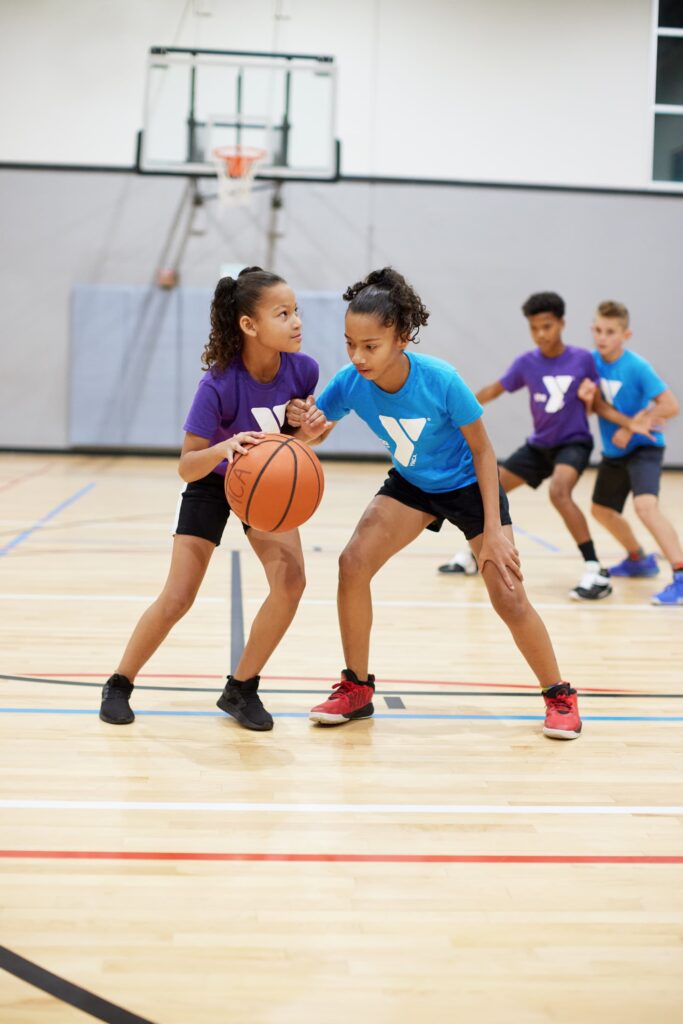 Two young girls are playing basketball on a court. One girl is wearing a blue shirt with a YMCA logo and black shorts. The other girl is wearing a purple shirt with a YMCA logo and black shorts. They are both holding a basketball and looking at each other.